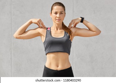 Waist up shot of good looking young female athlete, warms before jogging, keeps hands on shoulders, does sport exercises, dressed in tanktop, isolated over grey concrete wall. Fitness concept - Powered by Shutterstock