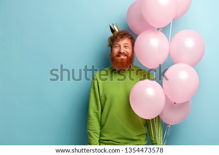 Waist up shot of handsome glad bearded man has birthday party, happy to recieve congratulations from friends, carries flying air balloons, has unforgettable festive event in life, stands indoor