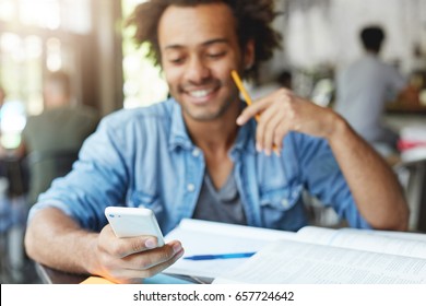 Waist Up Shot Of Happy Afro American College Student With Cute Smile Typing Text Message On Electronic Gadget, Sitting At Cafe Table With Textbooks. Selective Focus On Man's Hand Holding Cell Phone