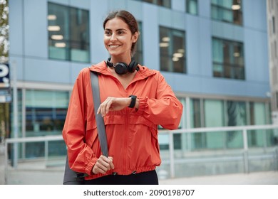 Waist up shot of happy brunette sporty woman dressed in anorak checks fitness results on smartwatch carries fitness mat waits for someone poses against modern city building. Healthy lifestyle - Shutterstock ID 2090198707