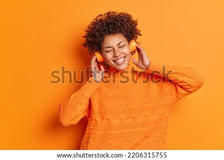 Waist up shot of happy curly haired woman keeps hands on stereo headphones listens her favorite songs keeps eyes closed dressed in casual jumper isolated over orange background. Hobby concept