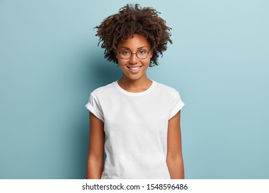Waist up shot of happy curly woman with toothy smile, wears optical glasses and casual solid white t shirt, expresses good emotions, enjoys nice day, isolated over blue background. Face expressions