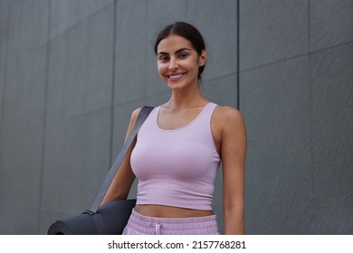 Waist up shot of happy motivated sporty woman has muscular arms good physical shape carries fitness mat going to have workout poses against grey background. Sports training and healthy lifestyle
