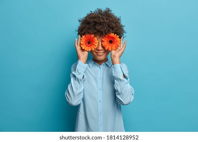 Waist up shot of happy woman seller of flower shop covers eyes with orange gerberas, dressed in stylish blue shirt and stands indoor against blue background. Tender female florist decorates room - Shutterstock ID 1814128952