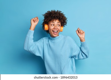 Waist Up Shot Of Joyful African American Woman Dances With Rhythm Of Music Wears Stereo Wireless Headphones Wears Casual Jumper Isolated Over Blue Background. Having Fun. Happy Female Meloman