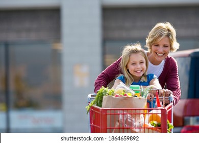 Waist Up Shot Of A Joyous Mother And Daughter Pushing A Shopping Trolley In A Supermarket Car Park With Copy Space.