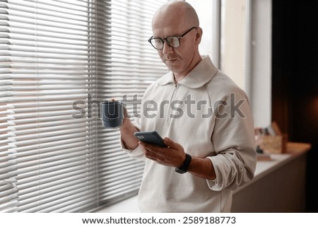 Waist up shot of middle aged man with buzz cut answering message on phone while drinking coffee by window during lunch break in office, copy space