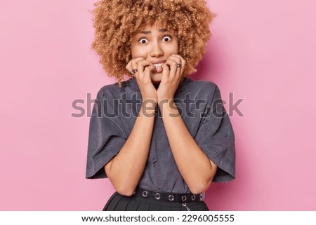 Waist up shot of nervous curly haired woman bite finger nails feels nervous looks anxious at camera wears grey t shirt isolated over pink background being confused and worried. Emotions concept