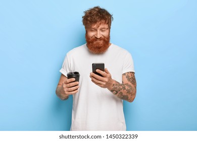 Waist up shot of pleased smiling red haired guy with thick beard, watches amused video online, holds mobile phone, drinks takeaway coffee, dressed in casual white t shirt, reads funny message - Shutterstock ID 1450322339