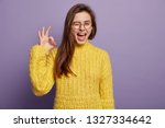 Waist up shot of pleased young woman says good job or well done, makes okay gesture, demonstrates symbol of approval and like, blinks eye, wears spectacles, knitted yellow jumper, stands indoor