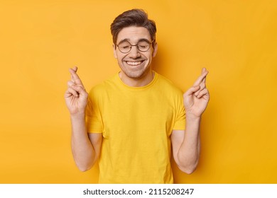 Waist Up Shot Of Positive Dark Haired Makes Cherished Wish Begs For Luck Hopes For Good Thinks Happen Wears Round Spectacles And Casual T Shirt Isolated On Yellow Background. Wish Desire Superstition