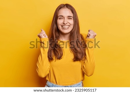 Waist up shot of positive European woman smiles gladfully clenches fists celebrates personal achievements or success dressed in casual jumper isolated over yellow background. Empowered triumph