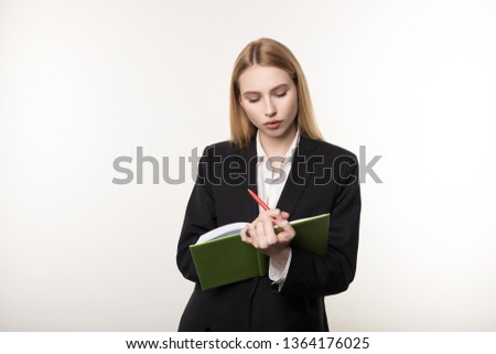 Waist up shot of serious blonde businesswoman in black suit that attentively listens to his business partner and takes notes in a notebook, isolated over white background