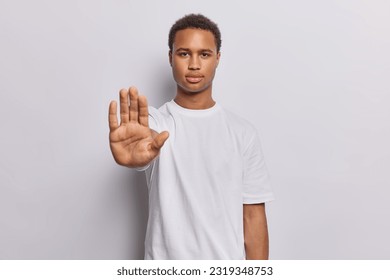 Waist up shot of serious dark skinned man raises his hand in firm stop gesture reminding us of importance of setting boundaries and taking pause dressed casually isolated over white background.