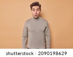 Waist up shot of shocked handsome European man stares bugged eyes at camera canot believe in astonishing news wears casual turtleneck isolated over beige background. Omg what horrible scene.