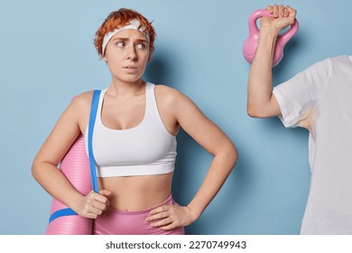 Waist up shot of sporty redhead woman in cropped white top and leggings carries karemat on shoulder looks at unknown person raising weight has no experience in training comes to gym for first time