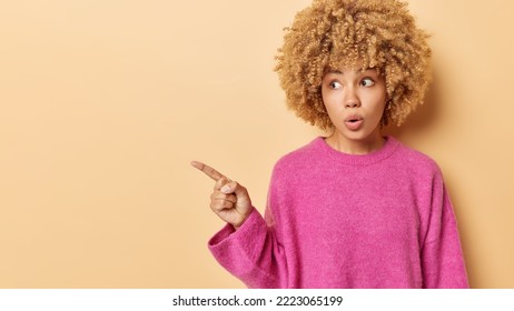 Waist up shot of surprised young woman has curly hair points finger left looks concerned and startled by something wears pink jumper shows product advertisement isolated over beige background