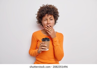 Waist up shot of tired sleepy curly brunette woman covers mouth and yawns drinks caffeine beverage to refresh wears casual orange jumper isolated over white background. People and tiredness concept