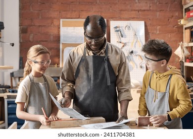 Waist up shot of two smiling children with senior carpenter looking at woodworking plans in workshop - Powered by Shutterstock