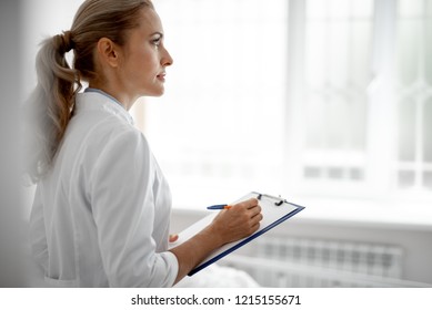 Waist up side view portrait of beautiful lady in white lab coat holding clipboard and pen while looking away with serious expression. Copy space on right side