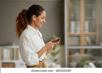 Waist up side view at smiling elegant businesswoman using wireless earphones and holding smartphone while standing at home or in office, copy space