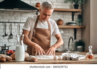 Waist Up Of Smiling Adult Man Standing In A Brown Apron And Kneading The Dough At Home. Baking Concept. Copy Space