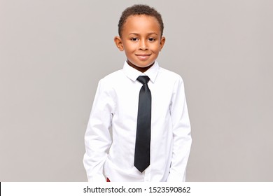 Waist Up Studio Shot Of Handsome Neat Dark Skinned Male Elementary Pupil Posing Isolated Dressed In White Clean Ironed Shirt And Black Elegant Tie, Ready For Going To School, Smiling At Camera