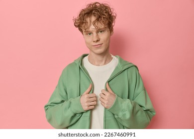 Waist up studio shot of a young curly red haired man looks and smiles at camera wears trendy clothes adjusts his green zipped hoodie over a pink background blank copy space for your advertising. - Powered by Shutterstock