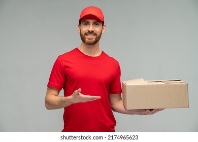 Waist Up View Portrait Of Male Courier In Red Uniform Handing Cardboard Parcel To Client, Carrying Parcel With Goods Ordered Online. Indoor Studio Shot Isolated On Grey Background 