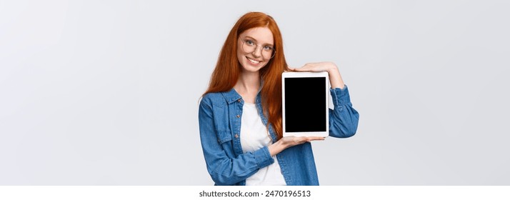 Waist-up attractive confident female with red hair, blue eyes in glasses, introduce application, presenting new app, holding digital tablet and showing gadget display, smiling recommend download. - Powered by Shutterstock