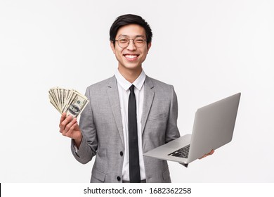 Waist-up Of Happy, Pleased Smiling Young Asian Man In Grey Suit Holding Dollars, Getting Rich, Using Laptop As Explaining How Invest In Financial Company, Winning Online Prize, White Background