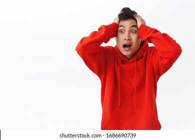 Waist-up portrait of alarmed asian man in red hoodie, panicking, grab head and running troubled, feel panic, gasping look sideways concerned, dont know what do, facing terrible situation