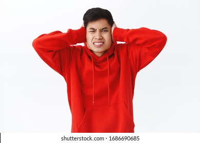 Waist-up Portrait Of Annoyed, Displeased Asian Man In Red Hoodie, Telling Neighbour Turn Volume Down, Shut Ears, Frowning And Grimacing Hear Annoying Loud Noise, Stand White Background