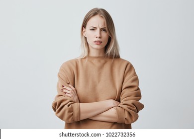 Waist-up portrait of beautiful girl with blonde straight hair frowning her face in displeasure, wearing loose long-sleeved sweater, keeping arms folded. Attractive young woman in closed posture. - Shutterstock ID 1022732356