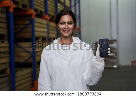 Waist-up portrait of a cheerful warehouse worker holding a smartphone in front of the camera