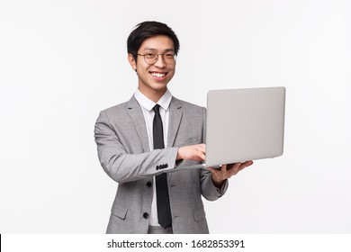 Waist-up Portrait Of Confident Successful Young Asian Man In Grey Suit, Standing Over White Background, Holding Laptop And Smiling At Camera, Working Remote, Using Computer To Show Presentation