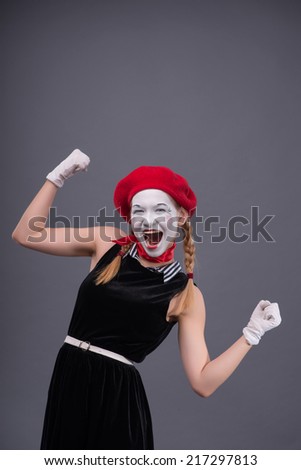 Waist-up portrait of female mime with funny face, red hat and red scarf looking at the camera smiling and showing that she has something done, isolated on grey background with copy place