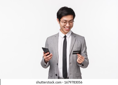 Waist-up Portrait Of Happy, Handsome Asian Male Entrepreneur, Office Worker In Suit, Holding Credit Card And Mobile Phone, Smiling, Easily Paying For Online Purchase Using Non-cash Payment Internet