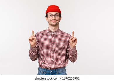 Waist-up portrait pleased cute bearded guy in red beanie and glasses, looking up sky enjoying spring fine day, found something cool, pointing and looking up, smiling at top advertisement - Powered by Shutterstock