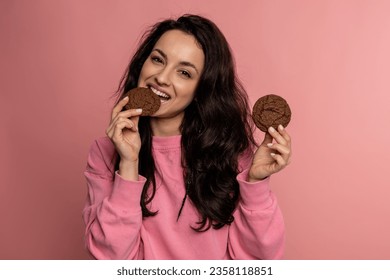 Waist-up portrait of a pleased dark-haired woman biting off a piece of oat biscuit while holding another in her hand. Sweet food concept