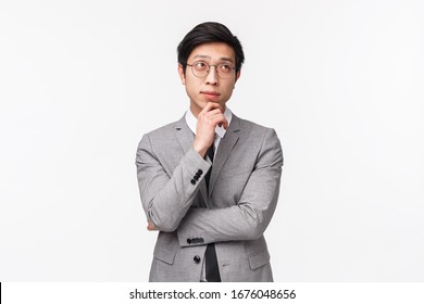 Waist-up Portrait Of Smart And Creative Young Asian Male Entrepreneur In Grey Suit, Making Plan, Hold Hand On Chin And Looking Up Thoughtful, Thinking Over Project, Standing White Background