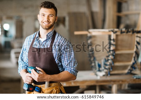 Waist-up portrait of smiling bearded craftsman with electric drill in hands standing in spacious workshop and looking at camera