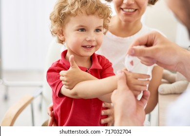 Waist-up portrait of smiling little boy showing thumb up while pediatrician putting bandage on injured finger, blurred background - Powered by Shutterstock