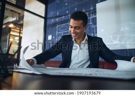 Waist-up portrait of a smiling pleased male employee studying a technical drawing at the desk