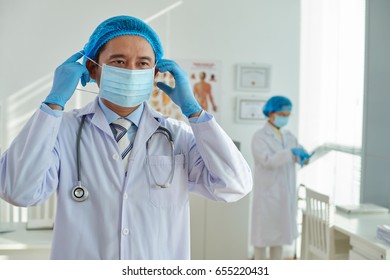 Waist-up Portrait Of Vietnamese Surgeon Putting On Medical Face Mask Before Operation, His Colleague Standing Behind Him