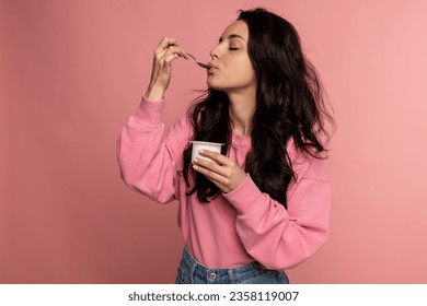 Waist-up portrait of a young woman snacking on the yogurt with a spoon from the plastic cup during the studio photo shoot. Healthy diet concept - Powered by Shutterstock