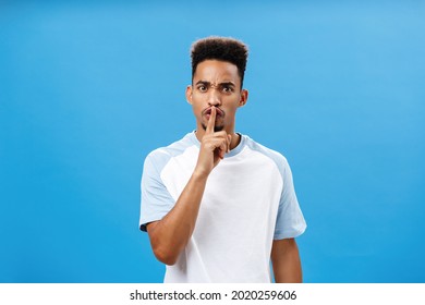 Waist-up shot of bothered and pissed good-looking african man with afro hairstyle frowning from irritation and annoyance demanding shut up keep secret and shushing at camera over blue wall