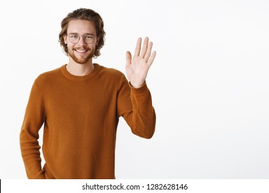 Waist-up shot of charming friendly man saying hi as waving with raised palm and smiling meeting new people smiling broadly welcoming members standing pleased and relaxed over gray background