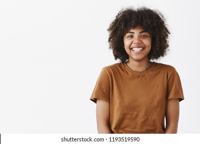 Waist-up shot of cute carefree friendly-looking African American teenage girl with afro hairstyle smiling broadly with shy and happy expression meeting new classmates over gray background - Shutterstock ID 1193519590