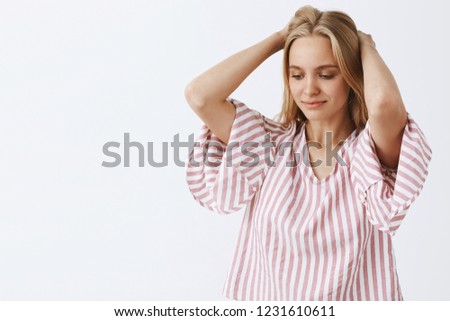 Waist-up shot of dreamy tender and feminine blonde caucasian woman in pink striped blouse holding hands behind head, gazing down with gentle slight smile thinking or imaging something